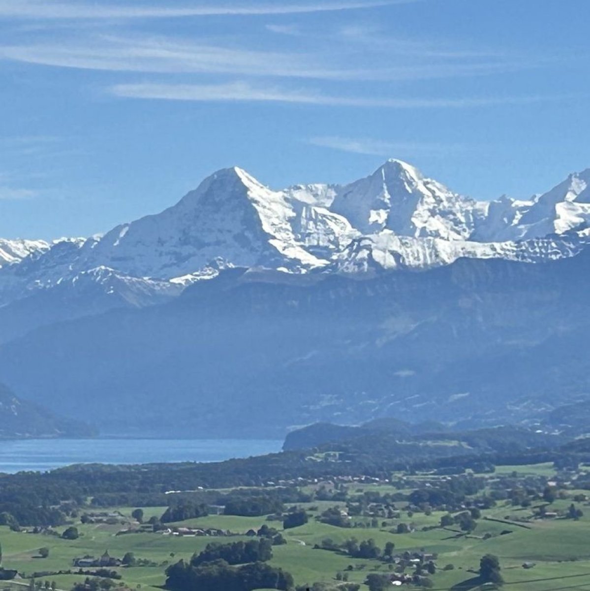 Schneebedeckte Berge über einem See und grünen Wiesen mit Häusern.